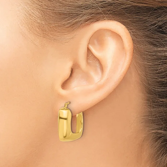 A person wearing a gold-toned chunky rectangular hoop earring with a polished finish, shown on their earlobe.