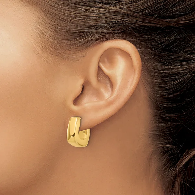 A person wearing a polished gold-toned rectangular hoop earring with a curved, chunky design on their earlobe.