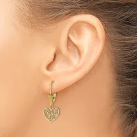 A woman wears a gold-toned heart-shaped dangle earring with decorative filigree detailing on a lever-back hook through her pierced earlobe.