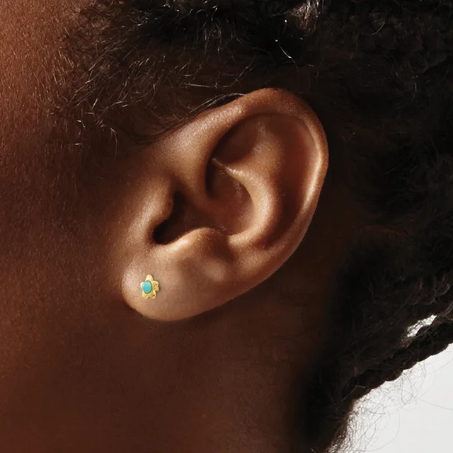 A small stud earring with a turquoise stone surrounded by decorative gold-colored metalwork worn on the earlobe of a person with dark skin.