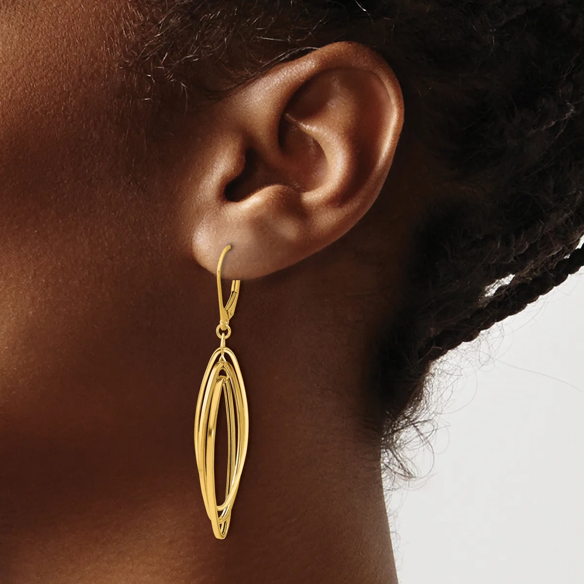 A gold-toned elongated oval drop earring with parallel ridged detailing hangs from a hook on a person's earlobe.