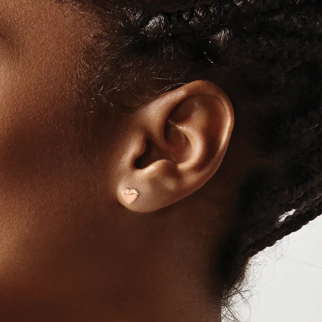 A small heart-shaped stud earring in silver or white metal worn on the earlobe of a person with medium brown skin tone and dark curly hair.