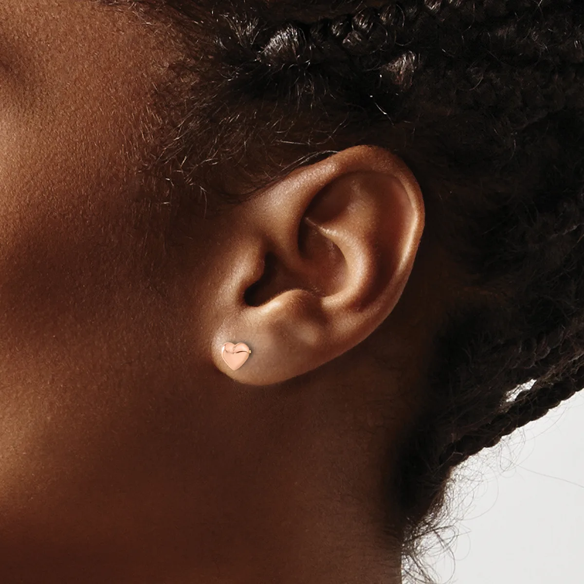 A small heart-shaped stud earring in silver or white metal worn on the earlobe of a person with medium brown skin tone and dark curly hair.