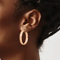 A woman wears rose gold hoop earrings with a rectangular cutout detail on each hoop, shown from the side profile against a neutral background.
