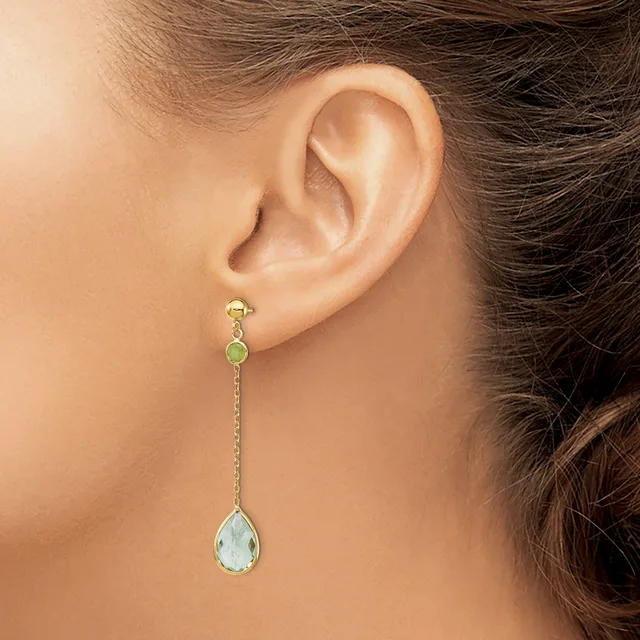 A woman wearing a long dangle earring with a green stone at the top, a textured gold chain, and a clear teardrop-shaped stone at the bottom.