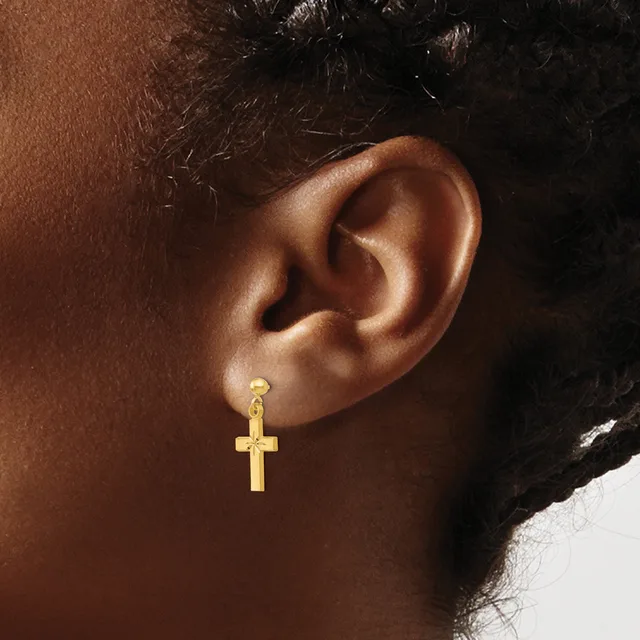 A gold cross dangle earring hangs from a pierced earlobe on a person with medium brown skin tone and black hair.