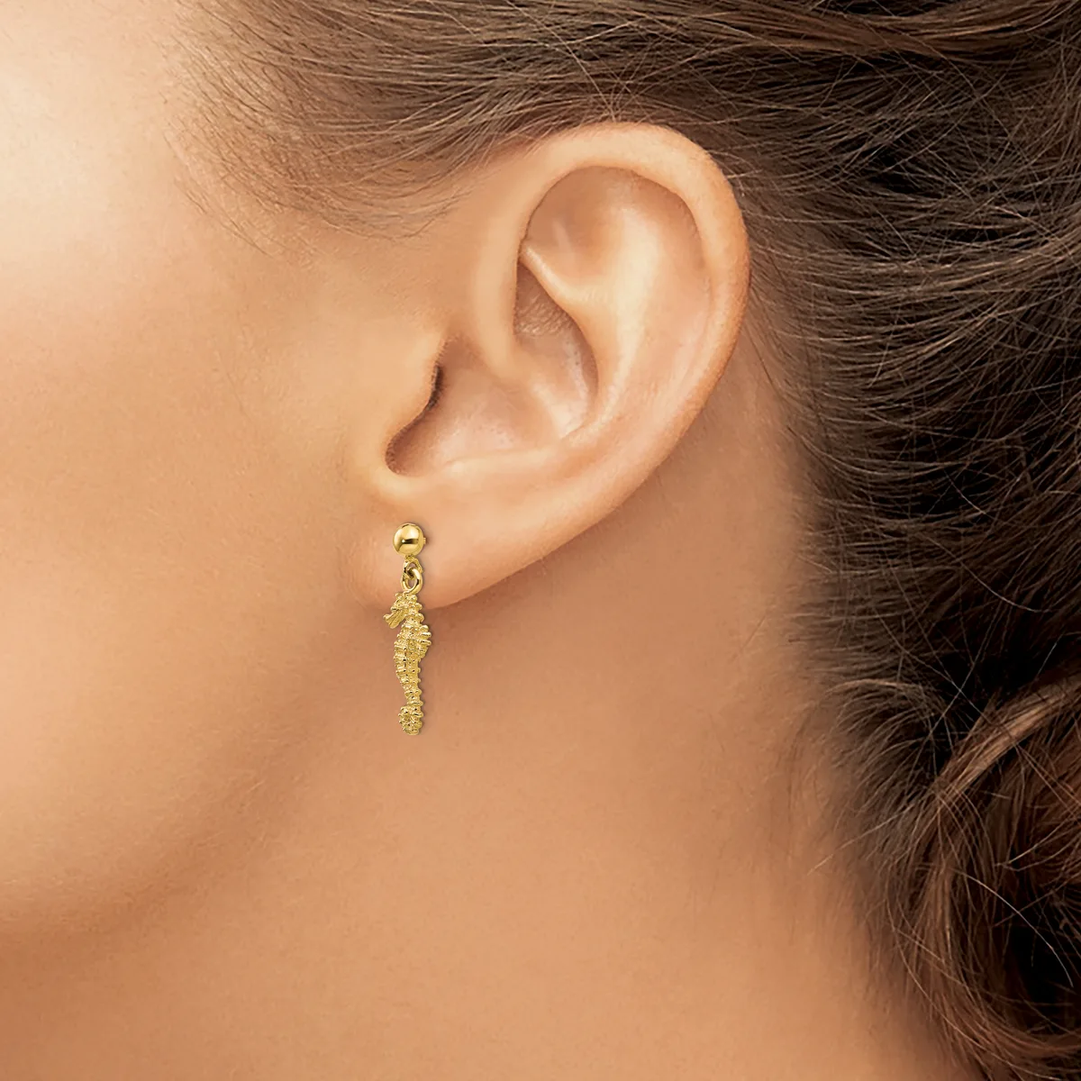A woman wearing a gold-toned dangle earring with a textured, elongated cylindrical design hanging from a small ball stud.