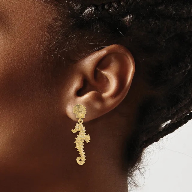 Gold seahorse-shaped dangling earring with textured finish shown on a model's ear with dark skin tone and black hair.