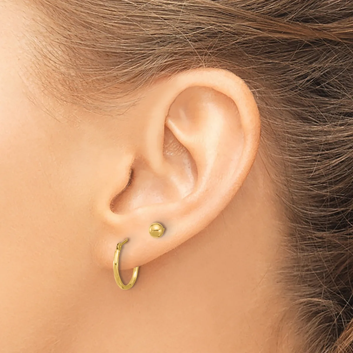 Close-up of a person's ear wearing two gold earrings: a small round stud and a small hoop.