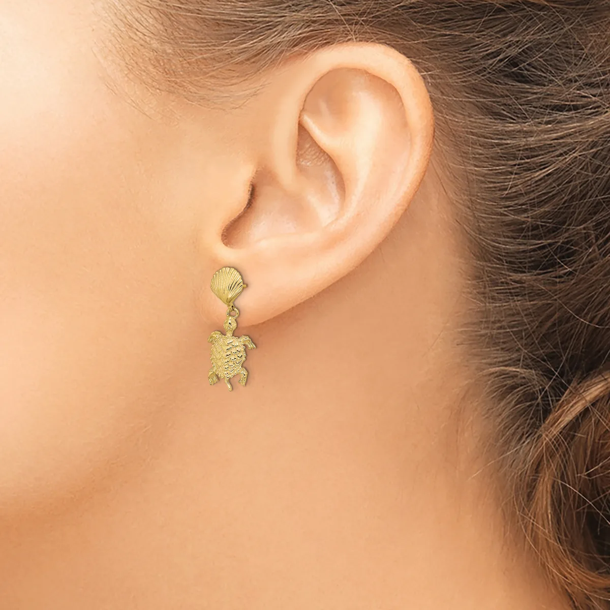 A person wearing a gold turtle-shaped dangle earring with a textured finish on a post and clutch back.