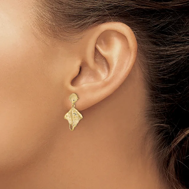 A close-up view of a person's ear wearing a gold textured drop earring with a leaf-like shape suspended from a small post.