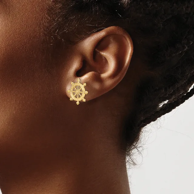 A close-up of a person's ear wearing a gold ship wheel-shaped earring with small beaded details along the bottom edge.