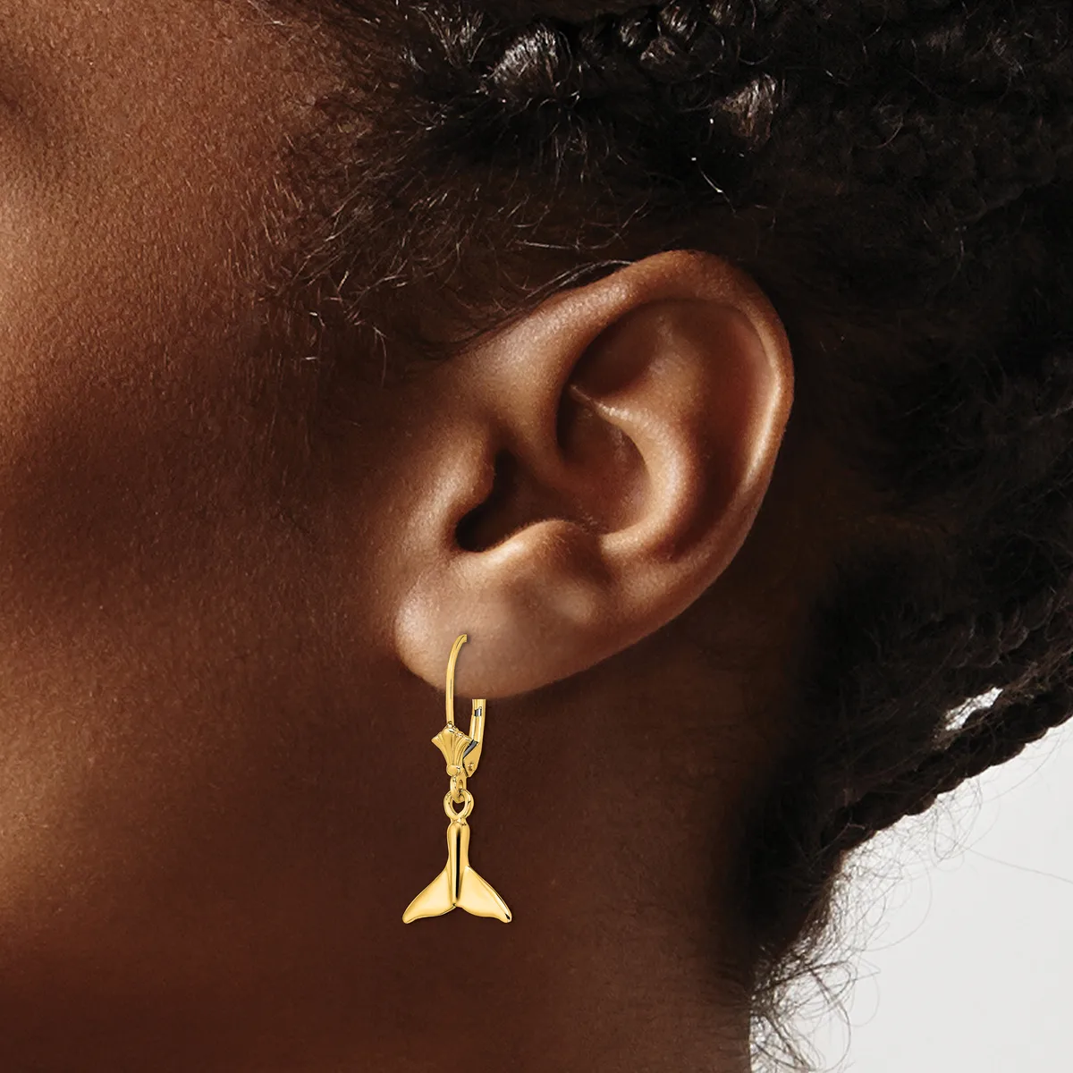 Gold whale tail charm earring hanging from a hook clasp on a person's earlobe.