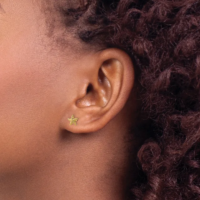 A small gold star-shaped stud earring worn in the earlobe of a person with deep burgundy curly hair and medium brown skin tone.