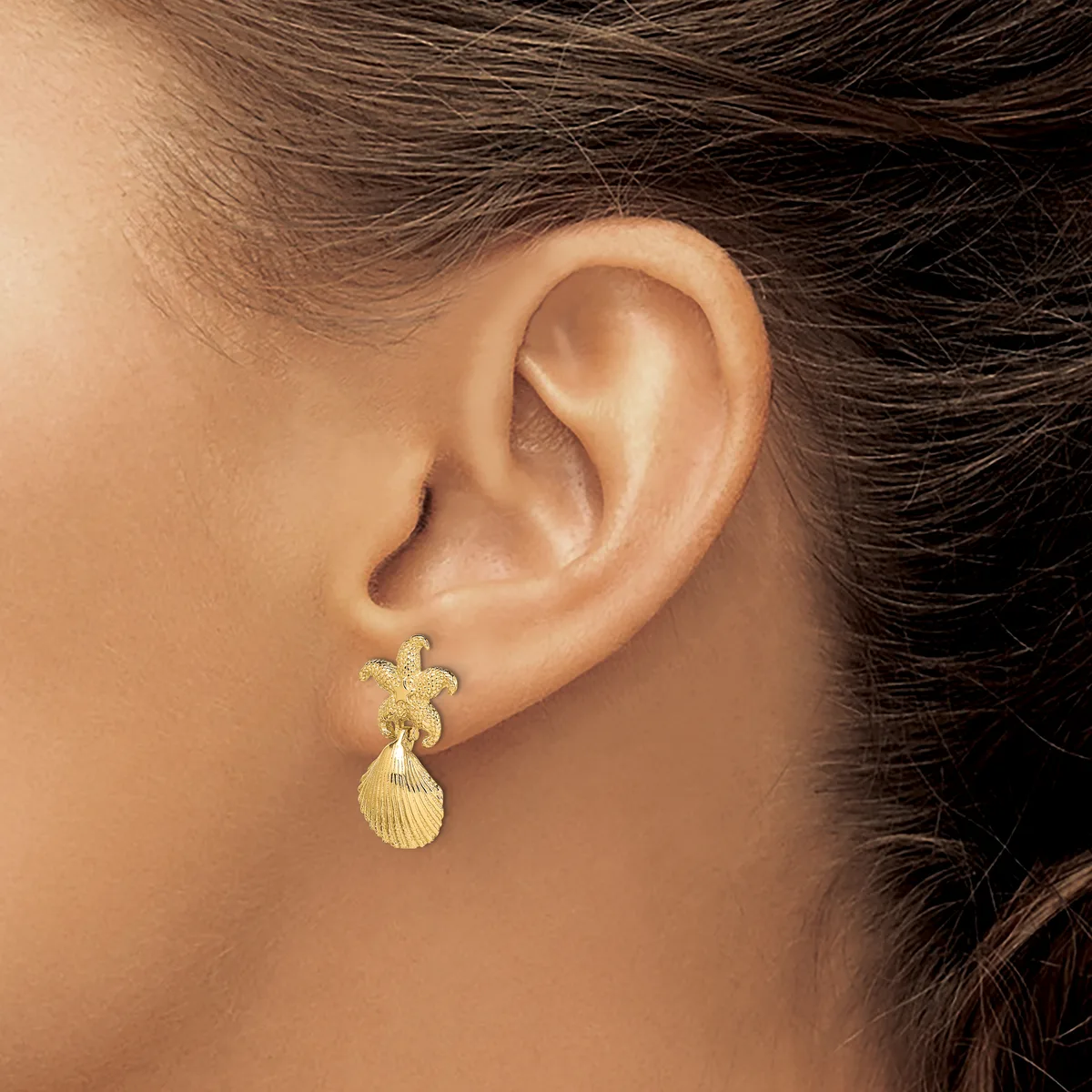 A person wearing a gold starfish and shell charm earring on their earlobe.