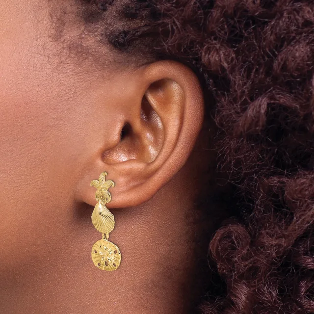A person wearing a gold drop earring with a shell-shaped top, textured middle section, and circular disc bottom, shown from the side profile.