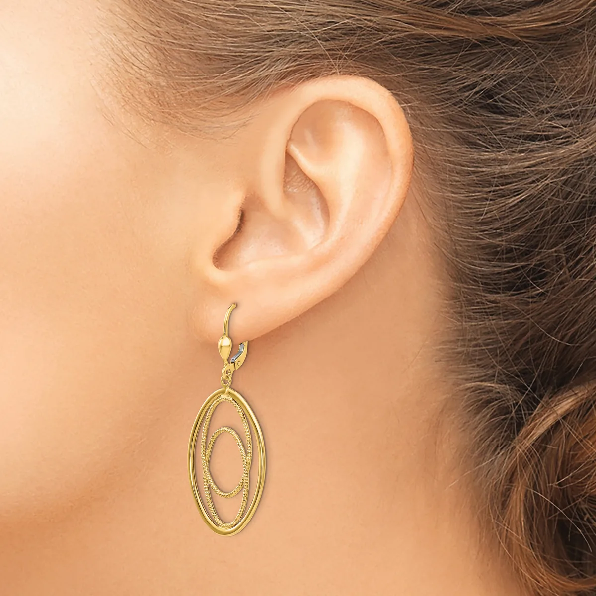 A woman wearing a gold-toned oval dangle earring with concentric oval designs, shown from the side view of her ear and neck.