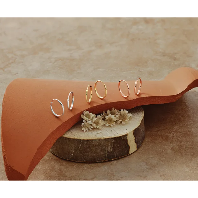 A tan leather cuff bracelet with small gold hoop earrings arranged on it, displayed on a wooden base with dried flowers.