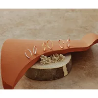 A tan leather cuff bracelet with small gold hoop earrings arranged on it, displayed on a wooden base with dried flowers.