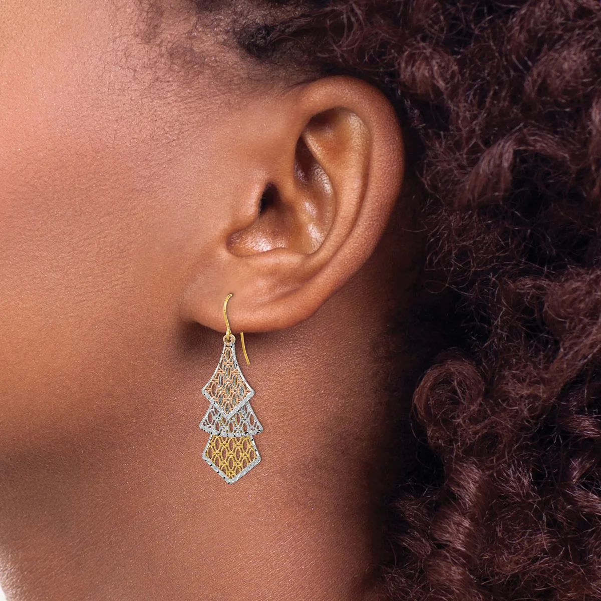 A person wearing a teardrop-shaped filigree earring with two-tone metalwork in gold and silver tones, suspended from a curved ear wire.