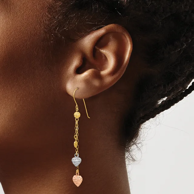 A person wearing gold drop earrings with a delicate chain, small gold bead, clear crystal heart, and rose gold heart charm hanging from a hook, shown from the side view of their ear.