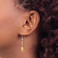A person wearing gold drop earrings with light blue gemstones and a gold teardrop charm, shown from the side view against dark curly hair.