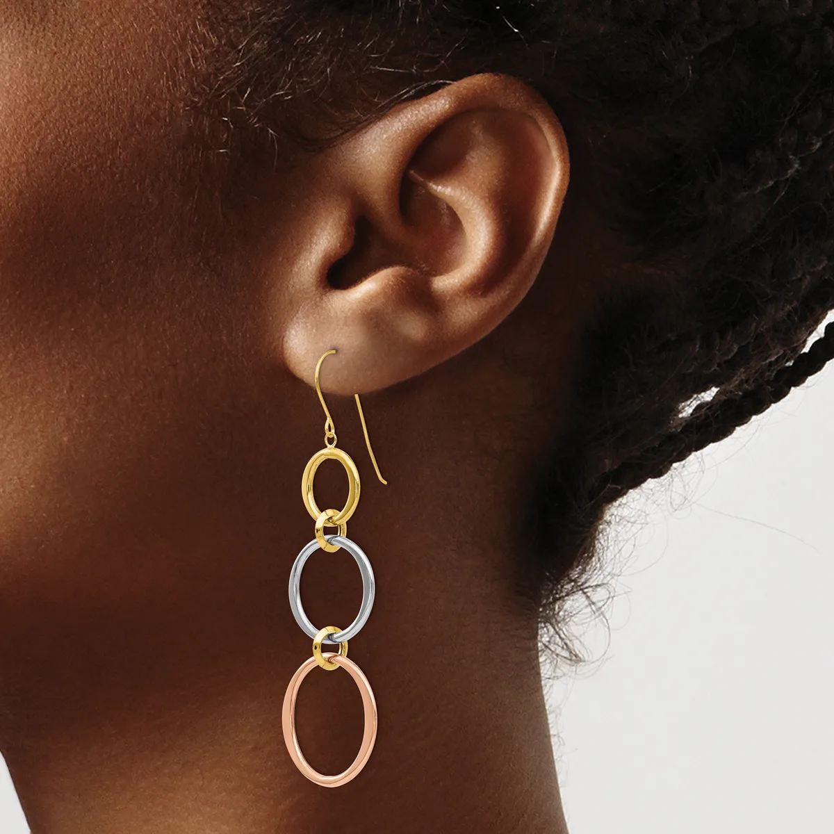 A person wearing drop earrings with three linked oval rings in gold, silver, and rose gold tones suspended from gold ear wires.