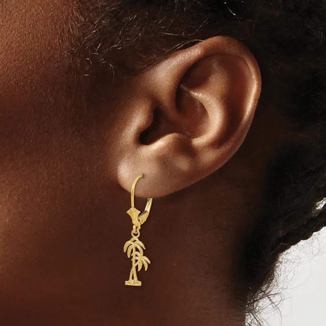 Gold hoop earring with a dangling palm tree charm worn on the earlobe of a person with brown skin.