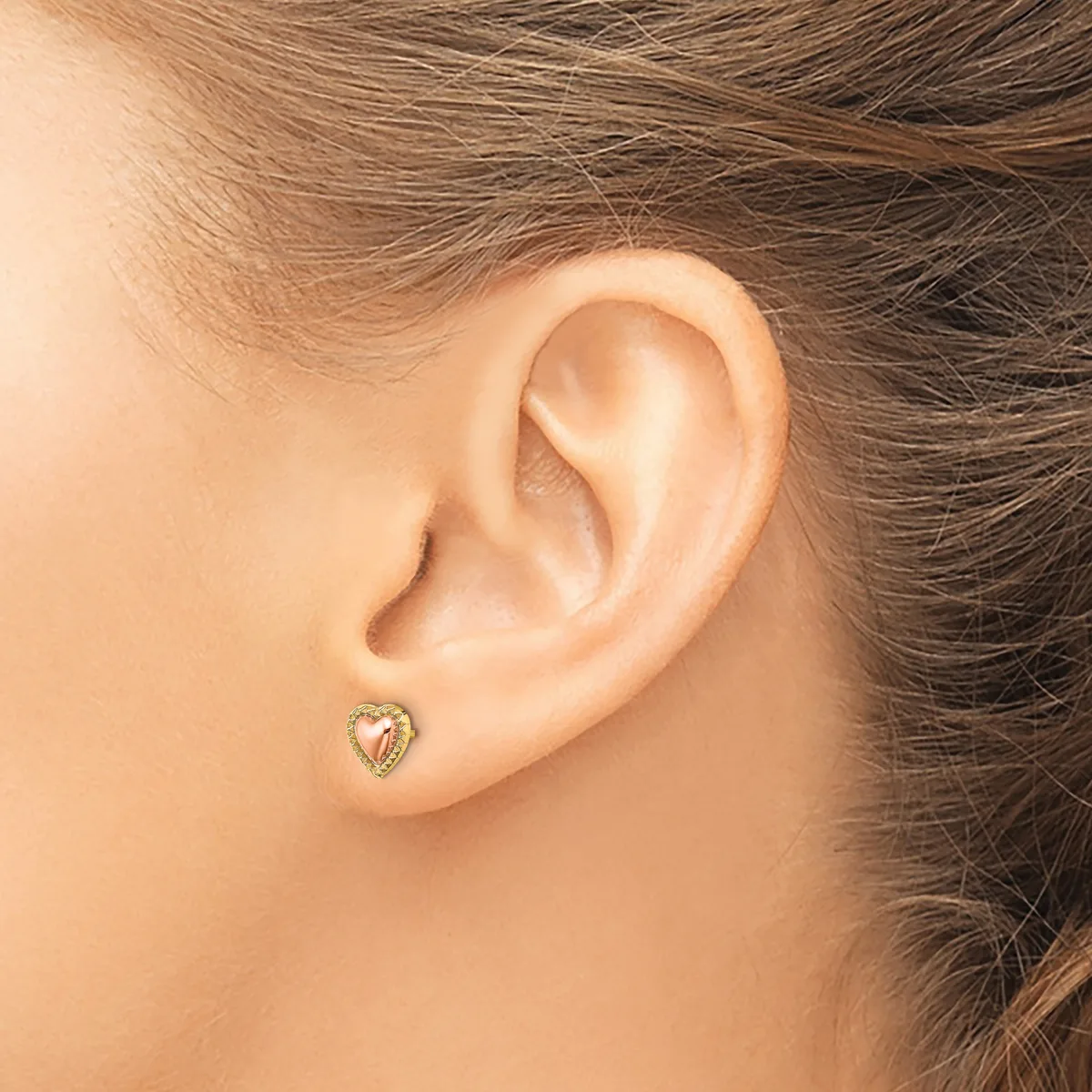 A small heart-shaped stud earring with a decorative border worn in the earlobe of a person with brown hair.