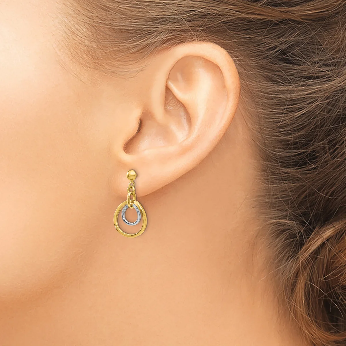 A woman wearing a two-tone dangling earring with nested oval loops in gold and silver, shown from the side view of her ear.