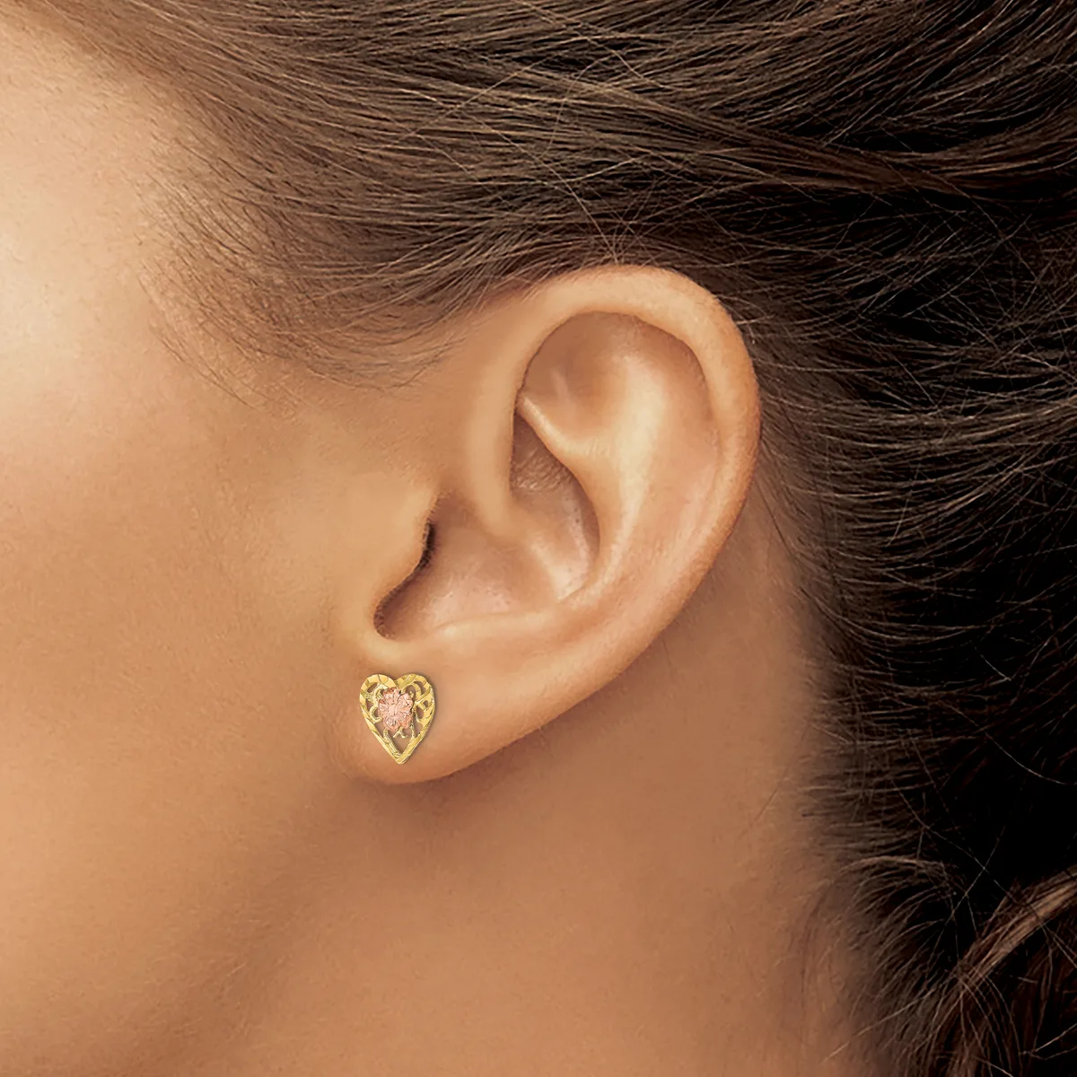 A person wearing a small gold heart-shaped stud earring with a filigree or textured surface, shown from the side view of their ear and face.