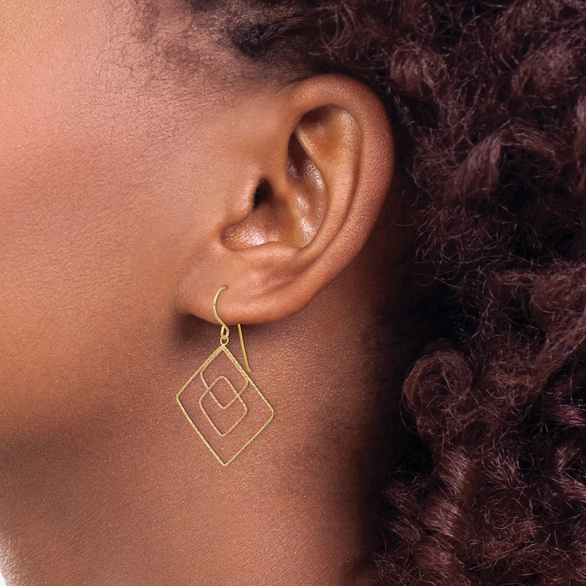 A person wearing a gold geometric diamond-shaped dangle earring with nested squares, shown from the side profile against their neck and curled dark hair.
