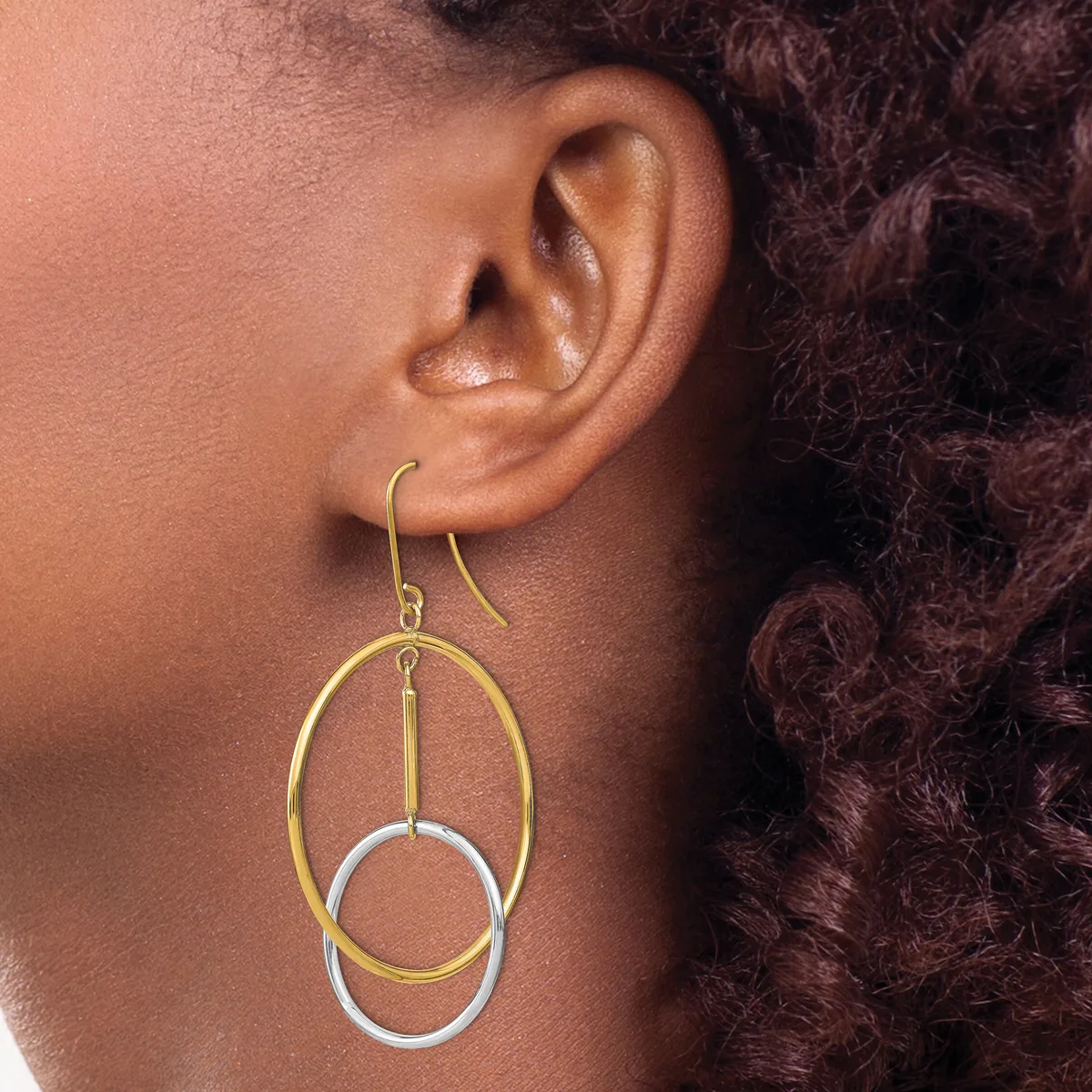 A person wearing gold and silver two-tone dangle earrings with nested oval and circular rings hanging from curved ear wires.