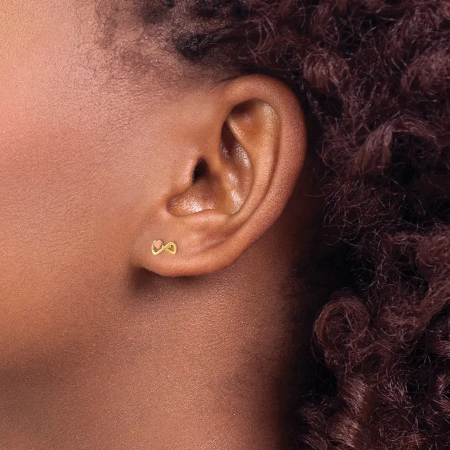 A person with dark curly hair wears a small gold stud earring shaped like an infinity symbol in their earlobe.