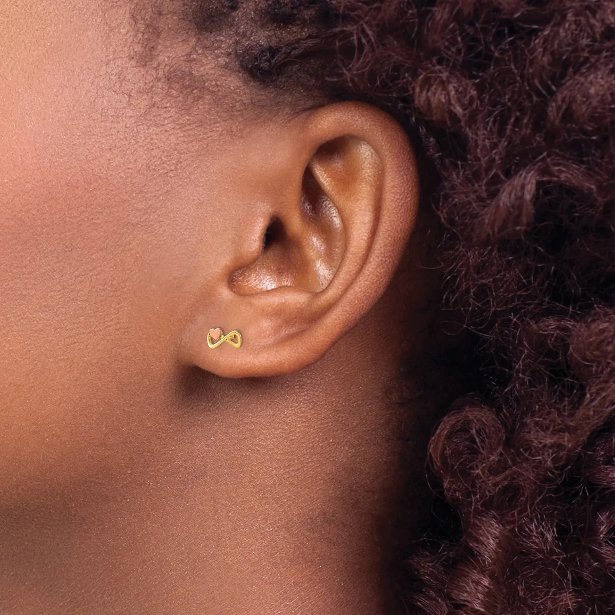 A person with dark curly hair wears a small gold stud earring shaped like an infinity symbol in their earlobe.