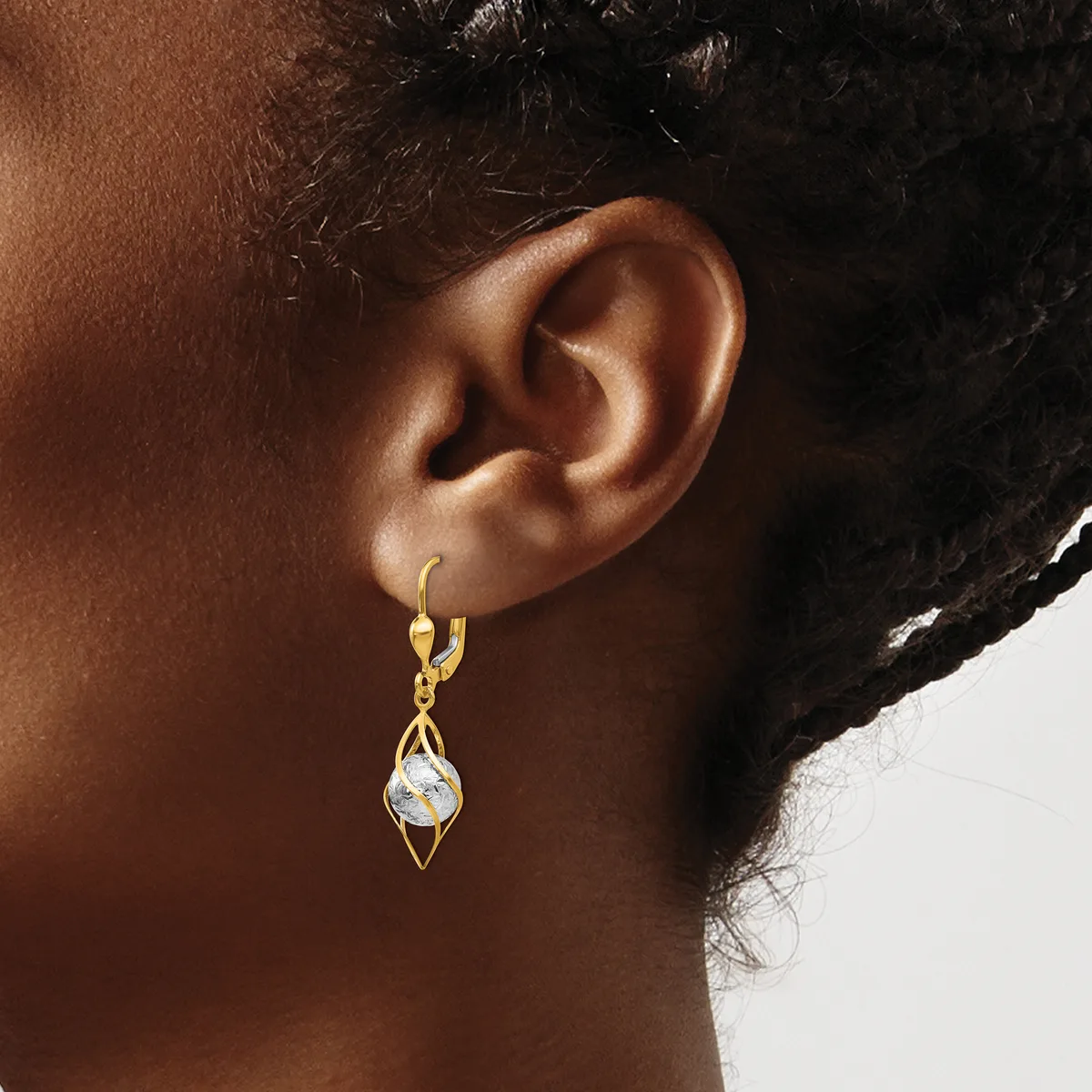 A person wearing a gold-toned drop earring with a diamond-shaped pendant and a clear stone, shown from the side view of their ear.