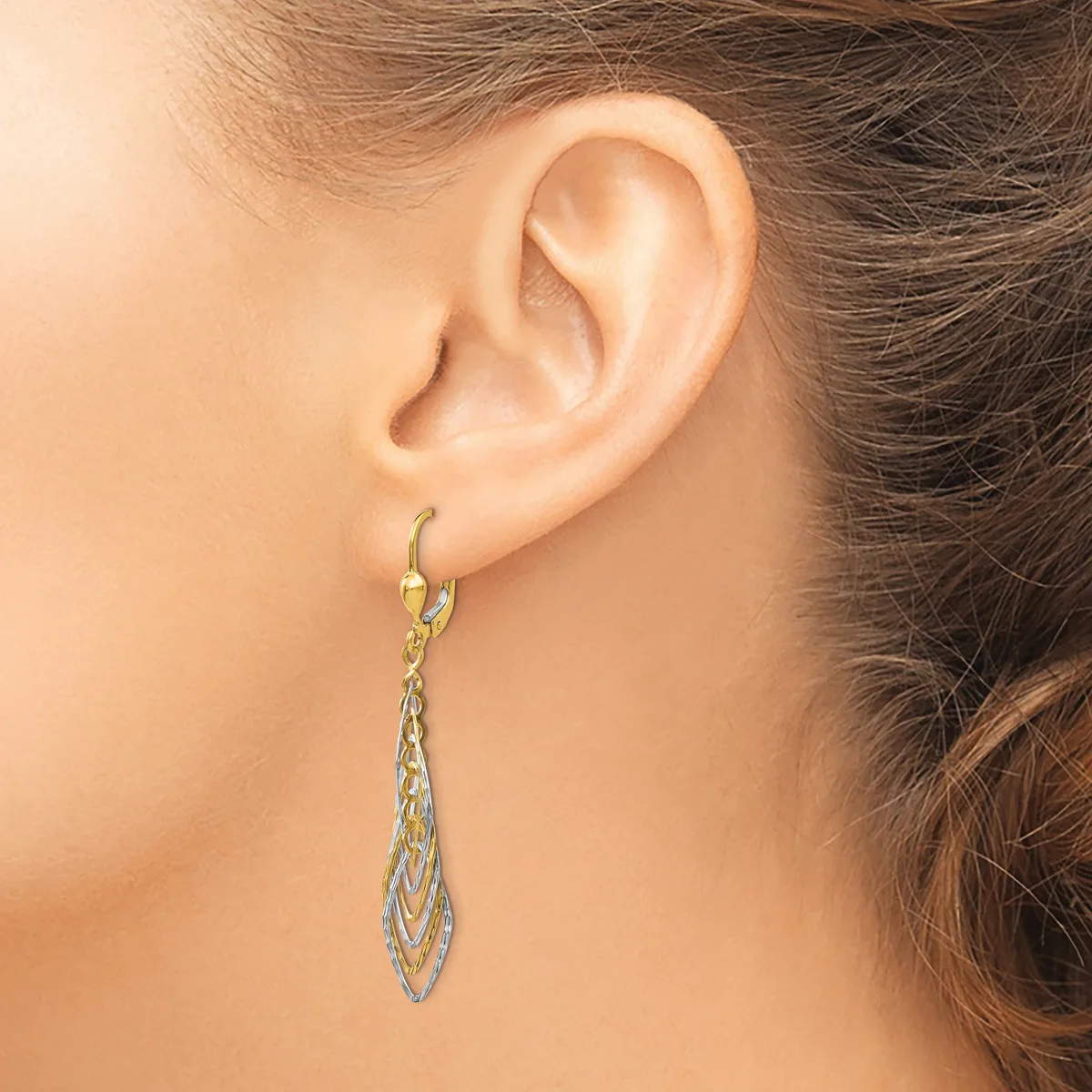 A woman wearing two-tone dangling earrings with textured teardrop pendants hangs from small hoop posts near her ear.
