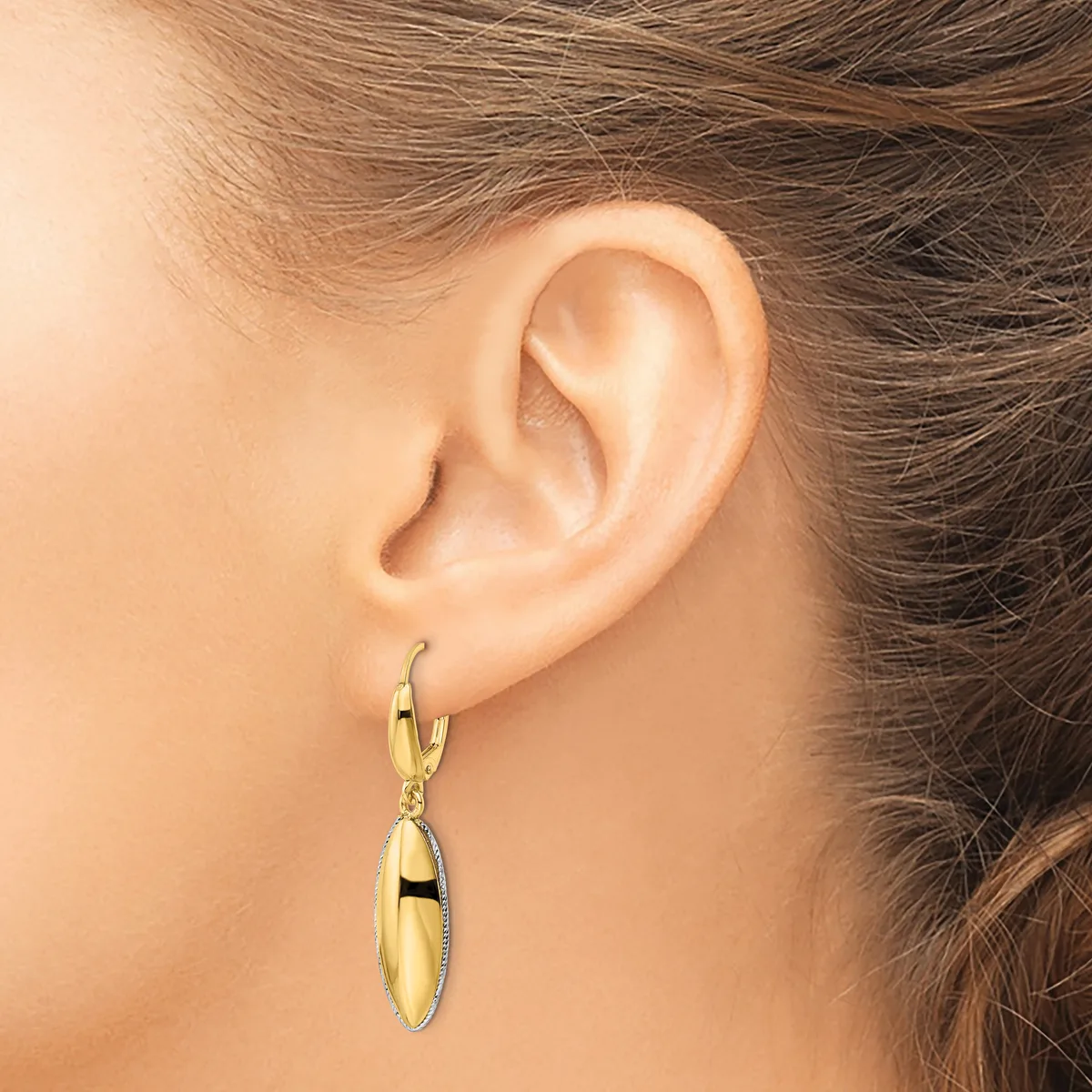 A person wearing a gold-toned oval dangle earring with a polished finish and textured border, shown from the side view against their ear.