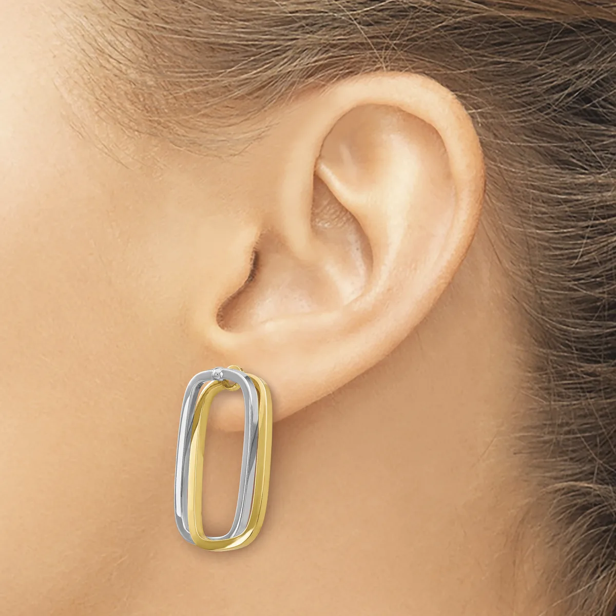 A person wearing a two-tone oval hoop earring in silver and gold metal, shown on their earlobe from the side.