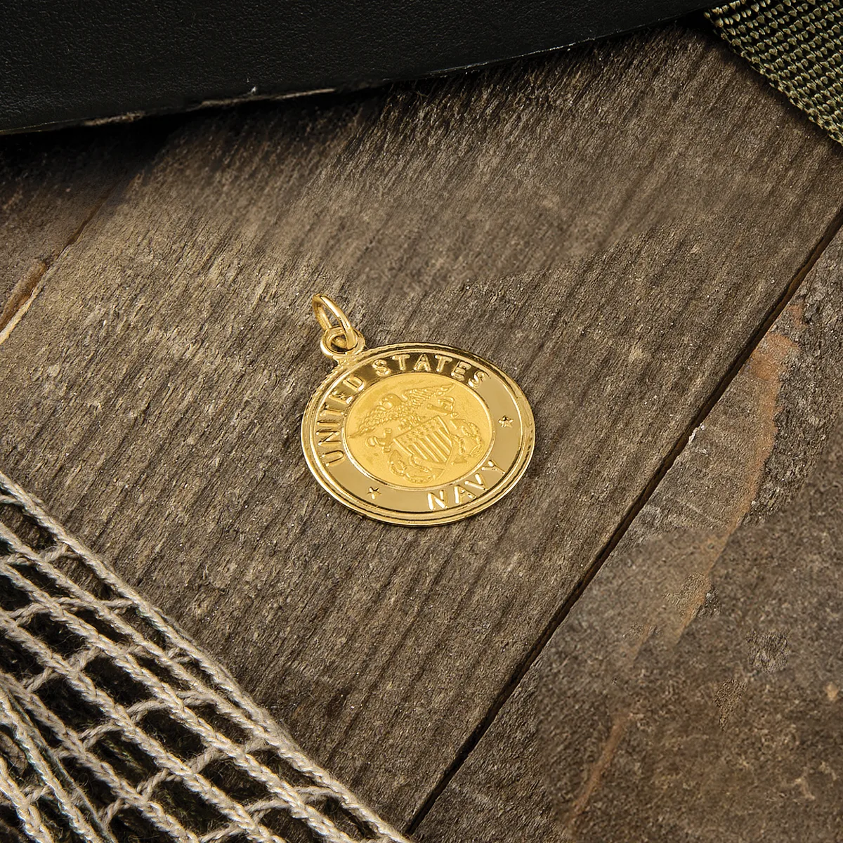 A gold-colored round pendant with a raised coin design in the center rests on weathered wood with fabric textiles nearby.