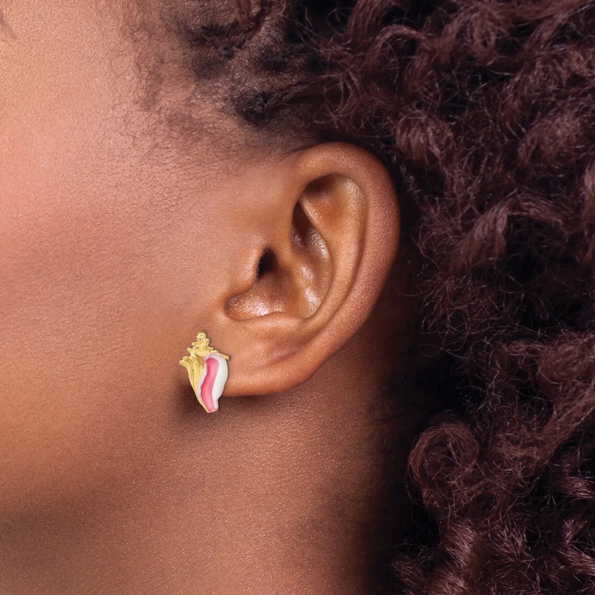 A person wearing a gold and pink enamel leaf-shaped stud earring in their pierced earlobe, shown in profile against their neck and curly dark hair.