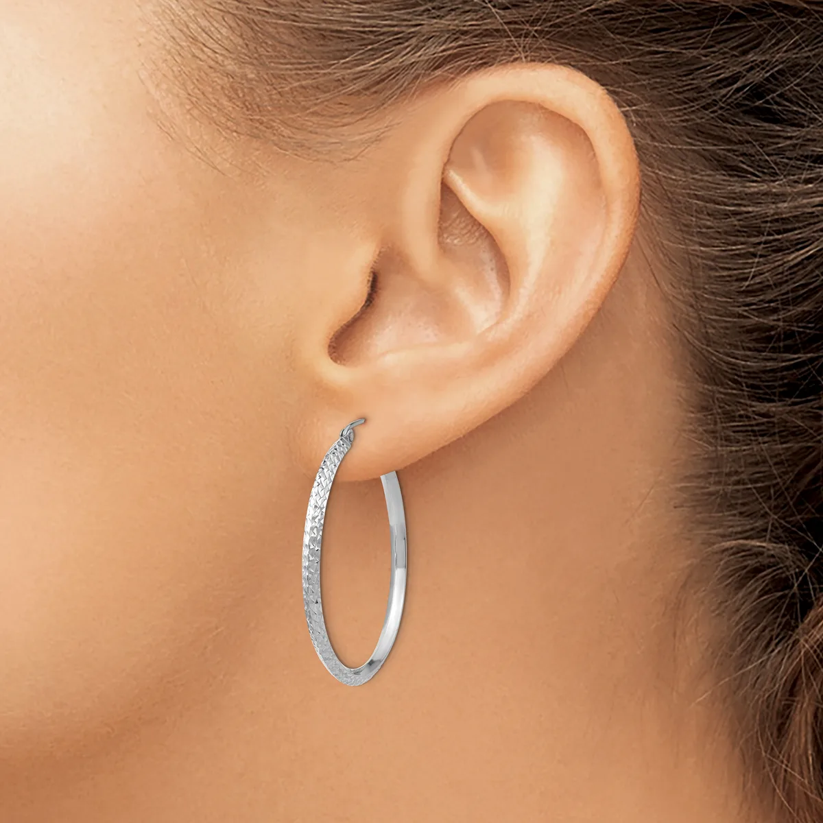 A woman wearing a pair of oval hoop earrings with a textured silver finish, shown from the side profile of her face and ear.