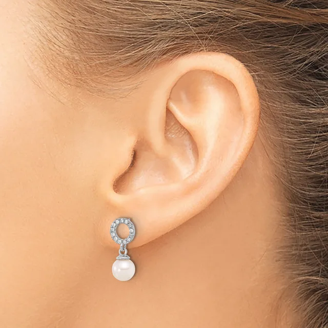 A close-up of a person's ear wearing a silver-toned drop earring with a circular rhinestone setting at the top and a white pearl pendant hanging below.