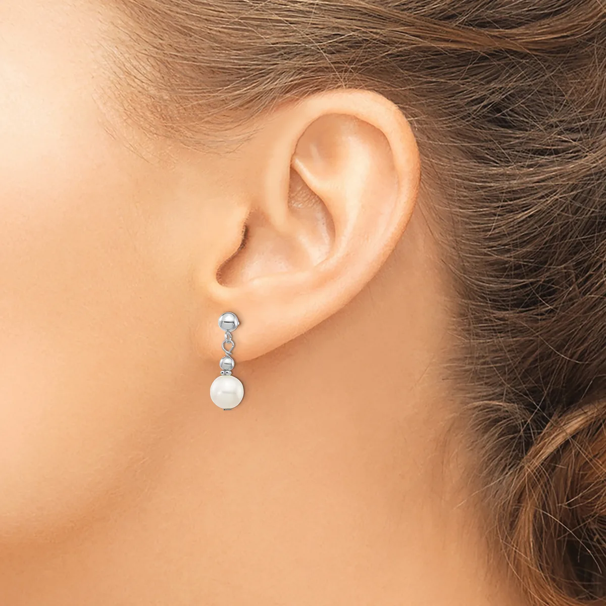 A woman wearing a dangling earring with a small blue stone at the top and a white pearl sphere below, connected by silver-toned metal links.
