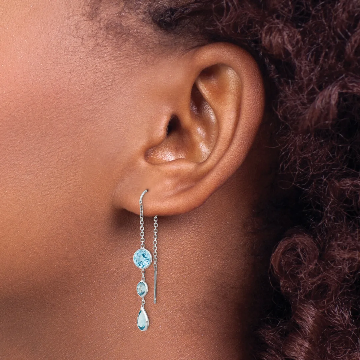 A person wearing a silver threader earring with light blue gemstones in graduated sizes hanging from a delicate chain through the ear.