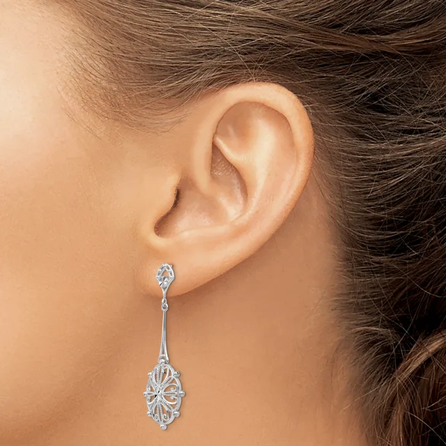 A woman wearing a long dangling silver earring with an oval filigree design and a small diamond stud at the top, shown from the side view of her ear and neck.