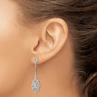 A woman wearing a long dangling silver earring with an oval filigree design and a small diamond stud at the top, shown from the side view of her ear and neck.