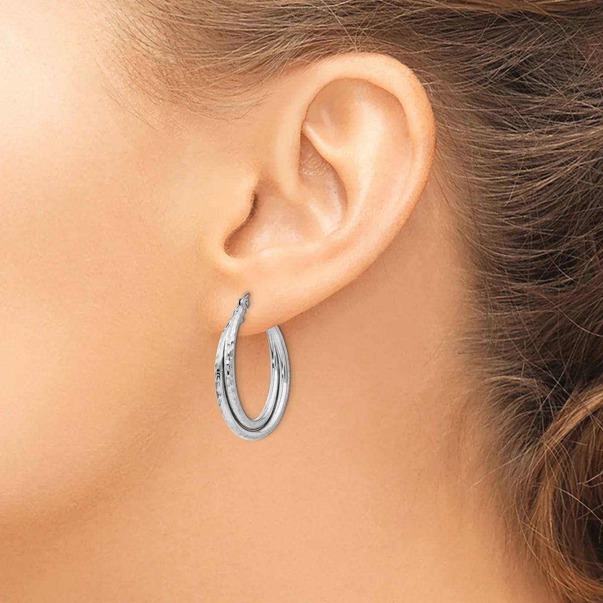 A woman wearing a pair of polished silver hoop earrings with a textured surface, shown from the side view near her ear.
