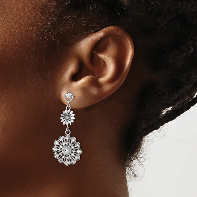 A woman wearing silver-toned drop earrings with small round stones at the top and larger ornate flower-shaped pendants below, shown from the side profile of her ear and neck.