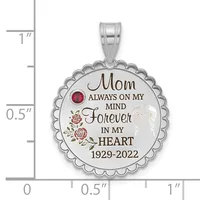 Silver scalloped circular memorial pendant with engraved text, roses, a red gemstone, and angel wing, shown next to a ruler measuring approximately one inch in diameter.